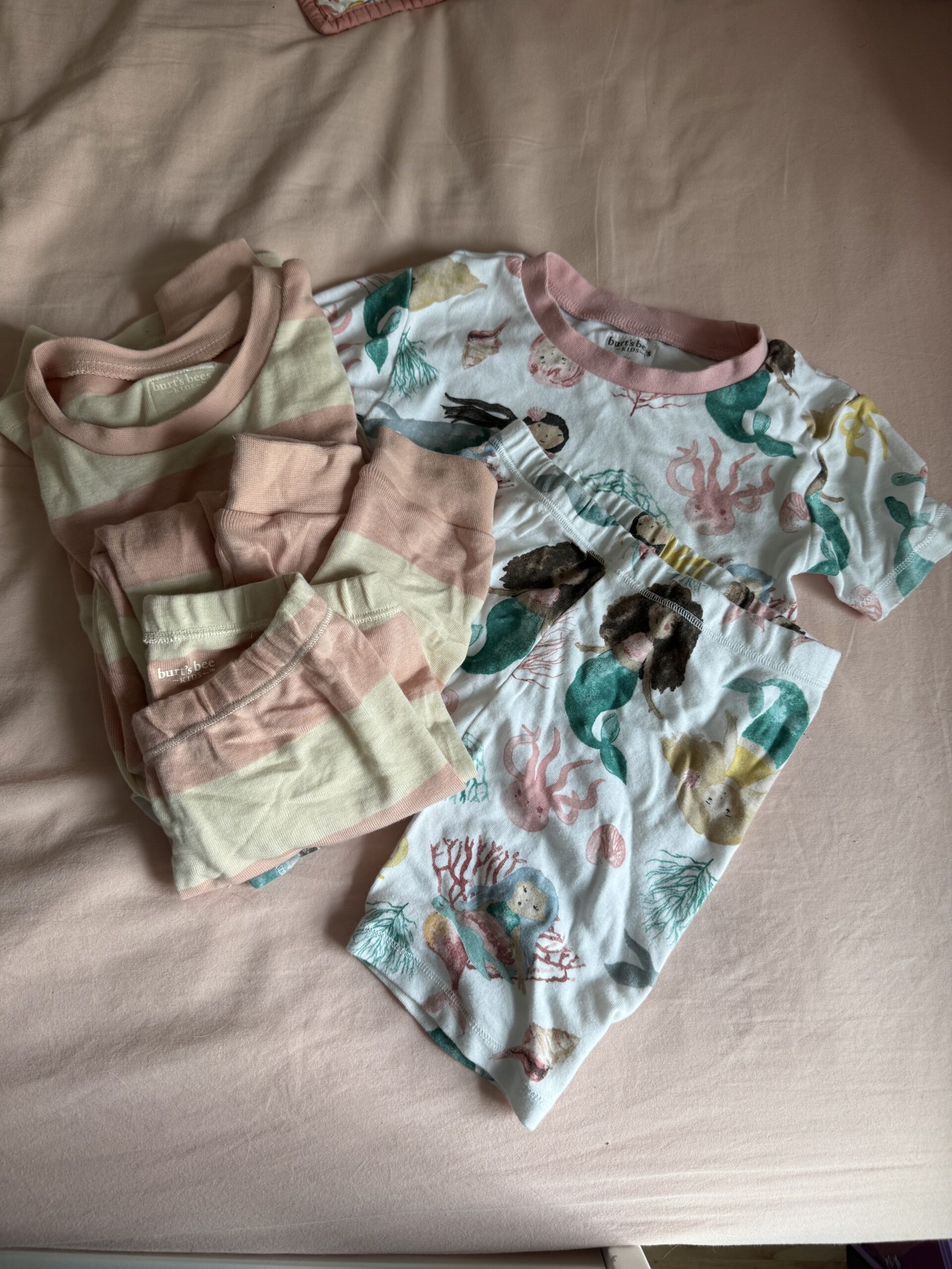 Two sets of children's pajamas are laid out on a pink bedspread; one set has pink and cream stripes, the other features a mermaid and ocean-themed print with pink trim.