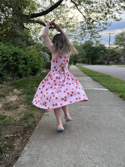 A young girl in a pink dress with red dots spins on a sidewalk near grass and trees on a cloudy day.