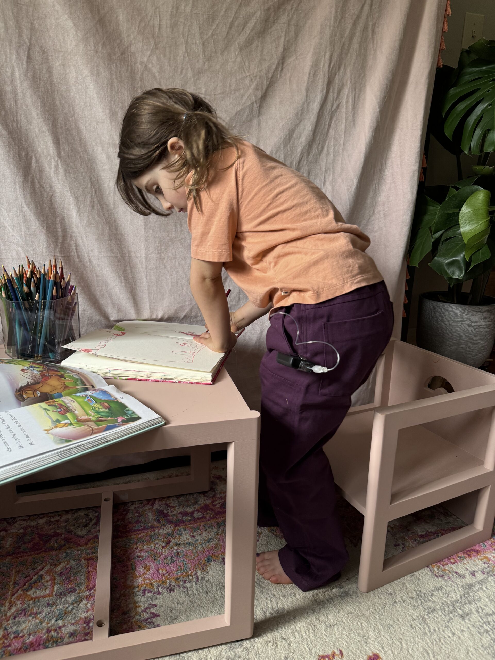 A young child stands at a small desk, drawing in an open notebook with colored pencils nearby, surrounded by books and a potted plant.