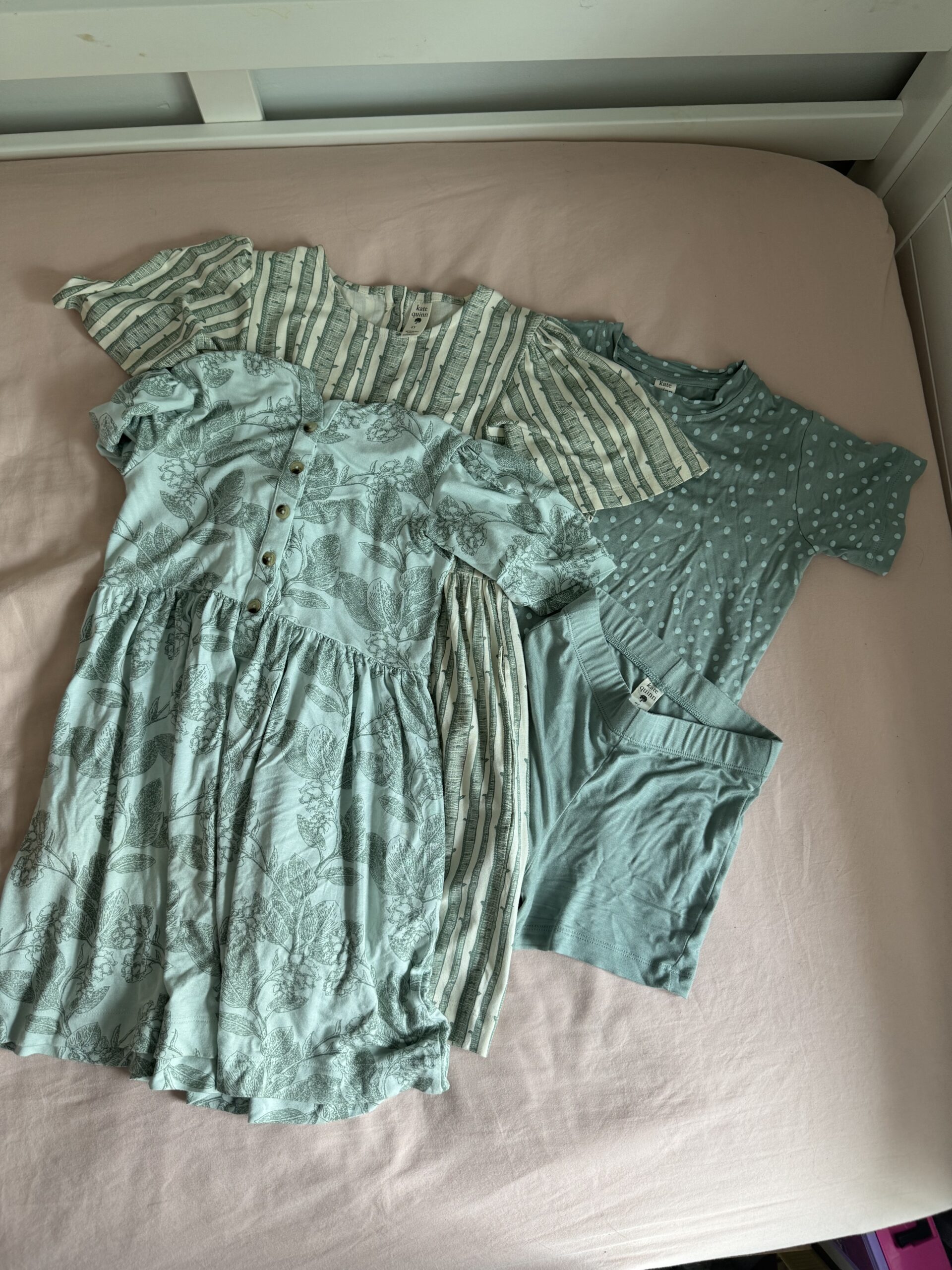 Four pieces of children’s clothing in shades of green are laid out on a pink bedsheet, including two dresses, a t-shirt, and a pair of shorts.