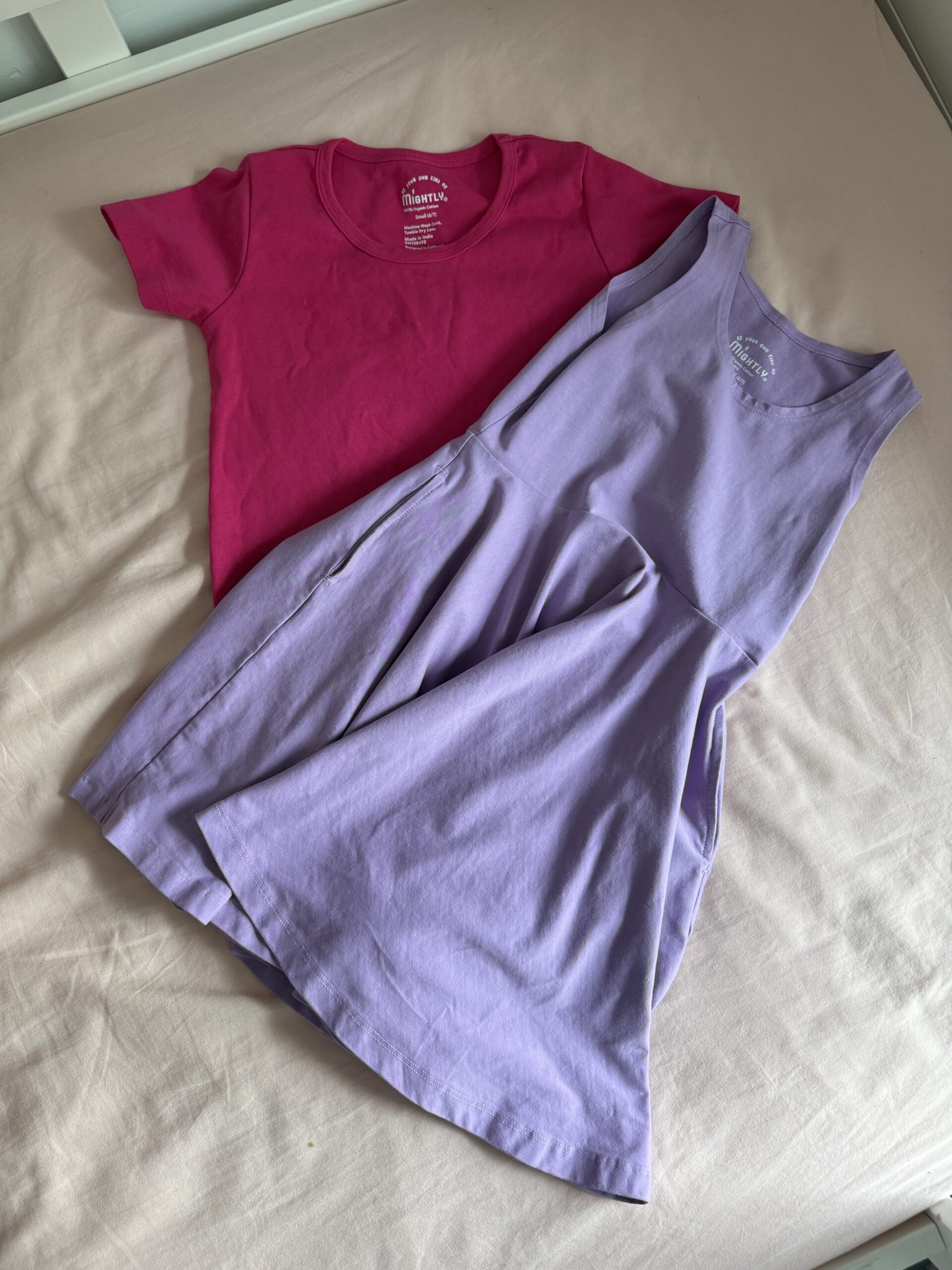 A pink short-sleeve t-shirt and a sleeveless lavender dress are laid out on a light beige surface.