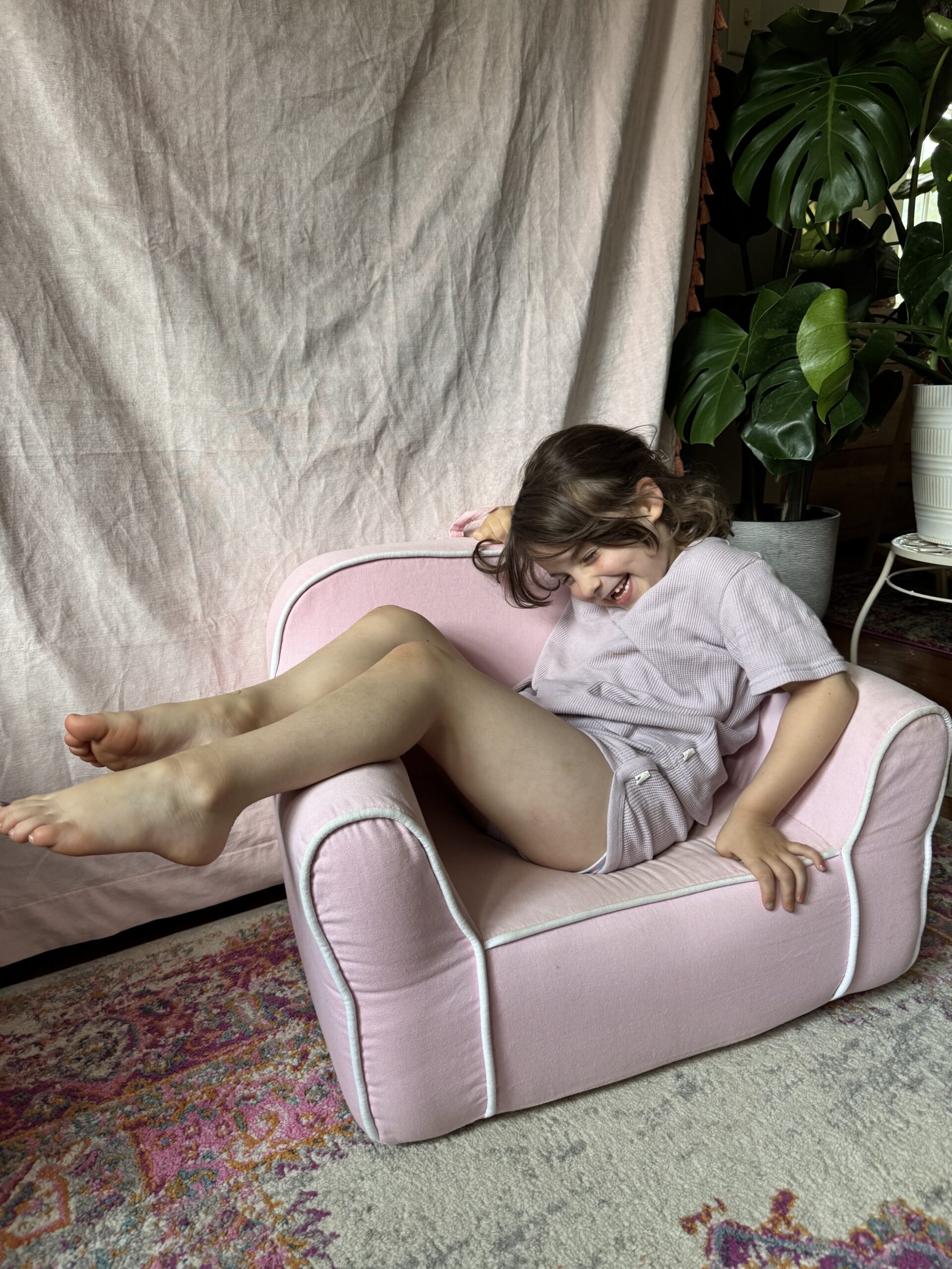 A young child sits sideways and smiles on a small pink armchair, with her legs resting over one arm. A plant and a pink fabric backdrop are visible in the background.