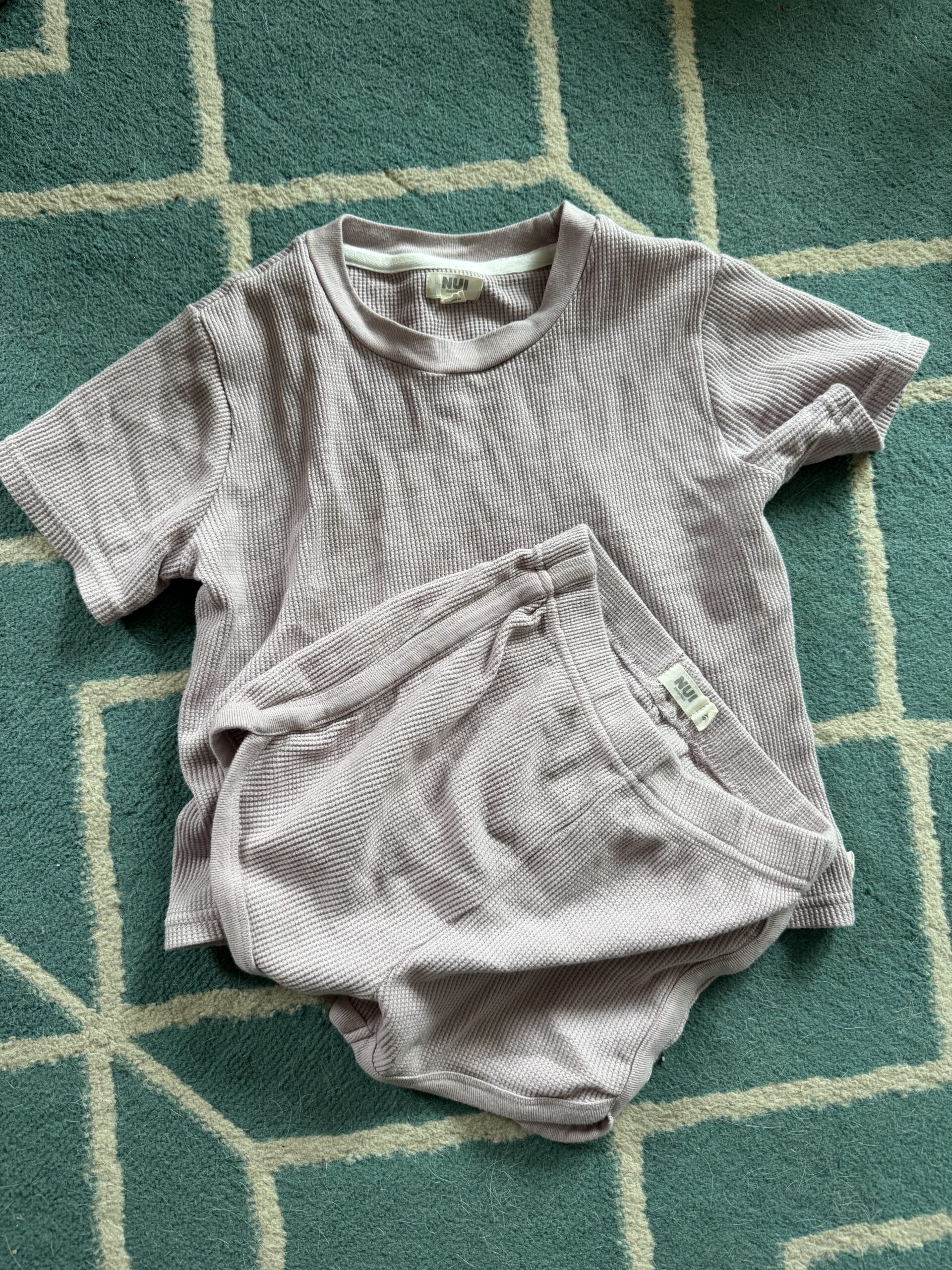 A short-sleeve ribbed lavender top and matching shorts set are laid out on a blue and white geometric-patterned rug.