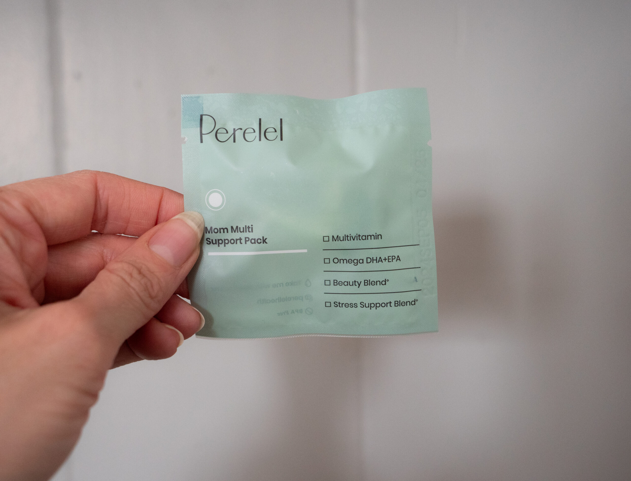 A hand holds a light green packet labeled "Perelel Mom Multi Support Pack" with checkboxes for Multivitamin, Omega, Beauty Blend, and Stress Support Blend against a plain background.
