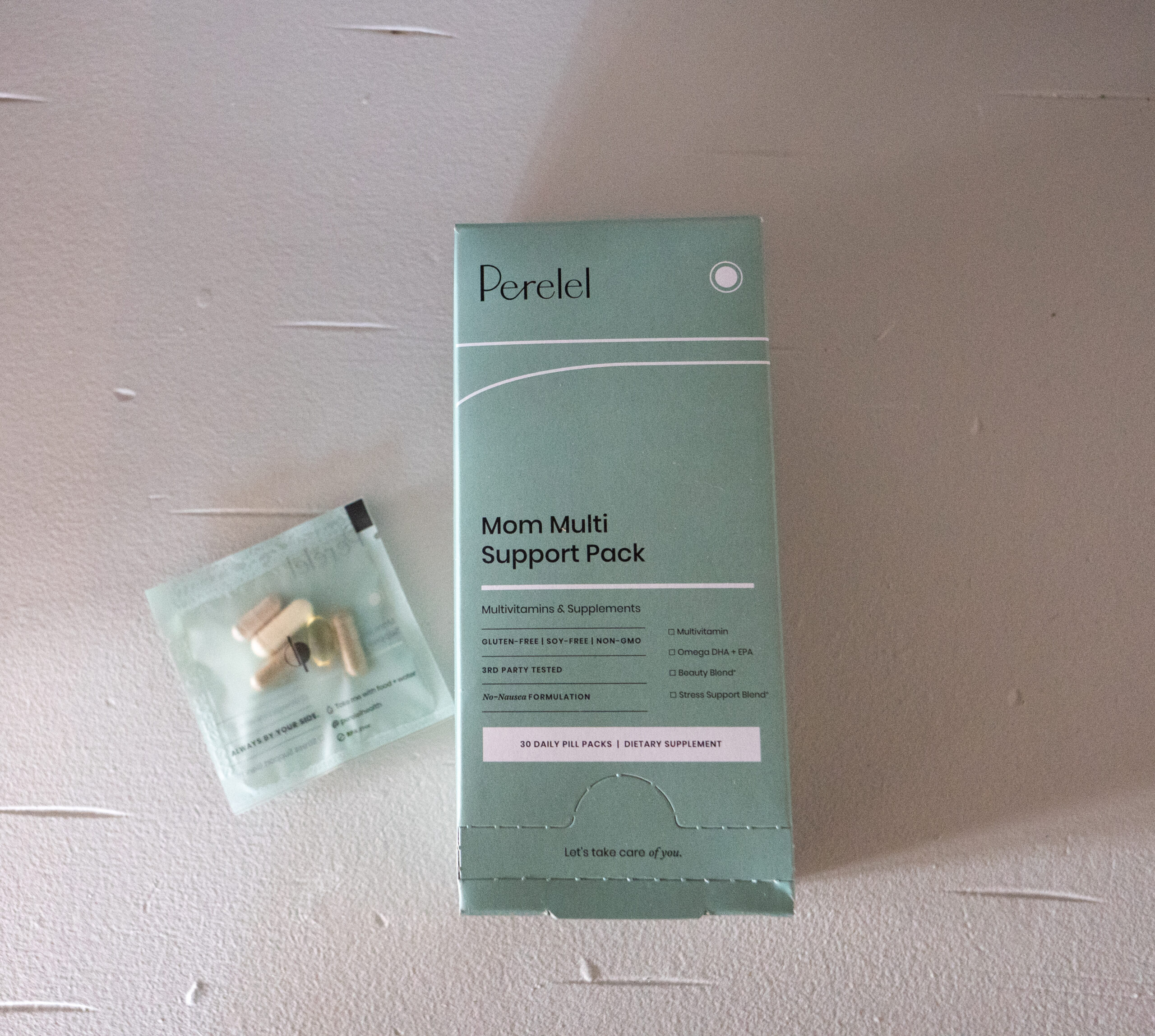 Box of "Mom Multi Support Pack" multivitamins with a small packet containing assorted pills, on a light surface.
