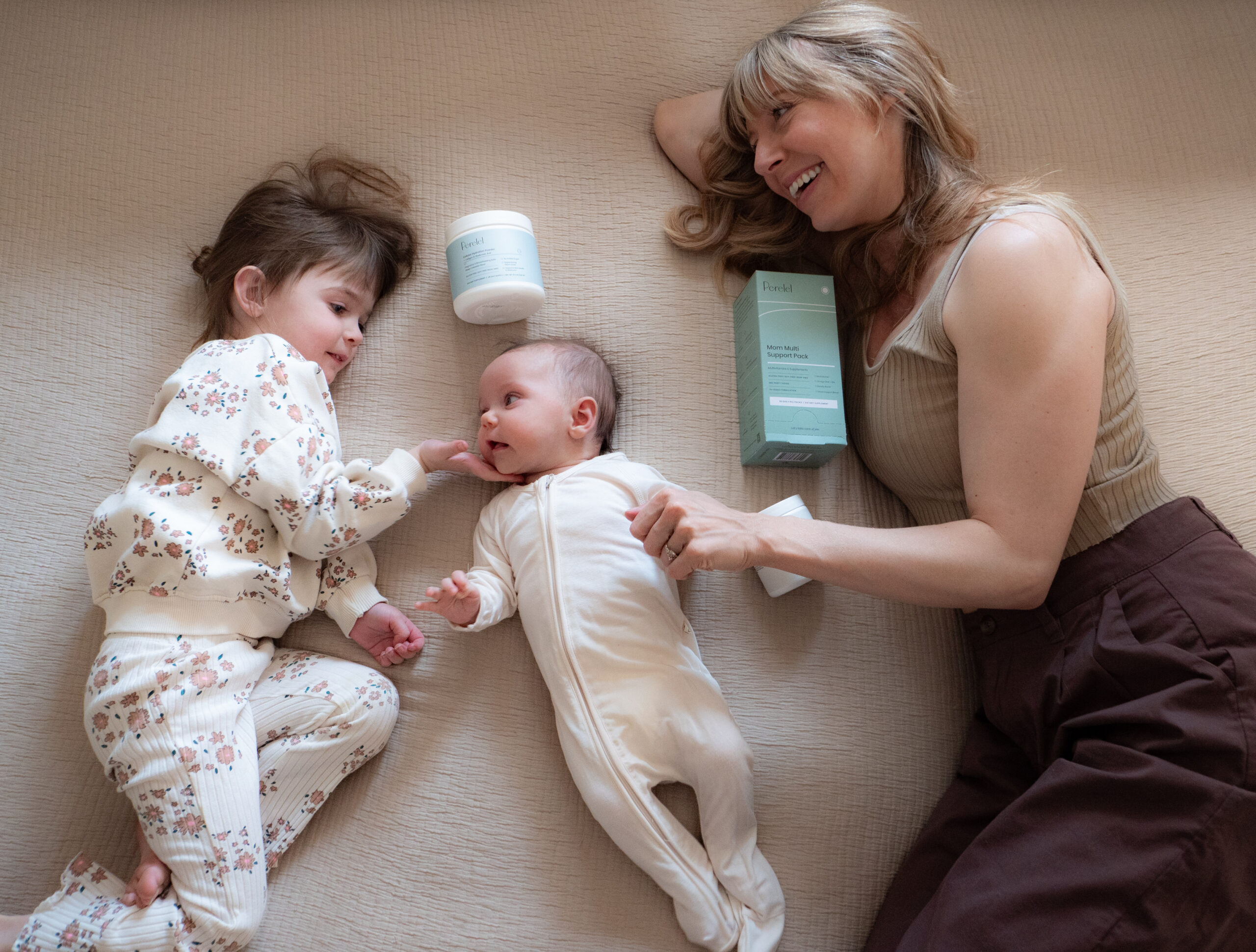 A woman lies on a bed with a baby and a young child. The woman is smiling, looking at the baby. There are two containers, one with a pump, nearby.