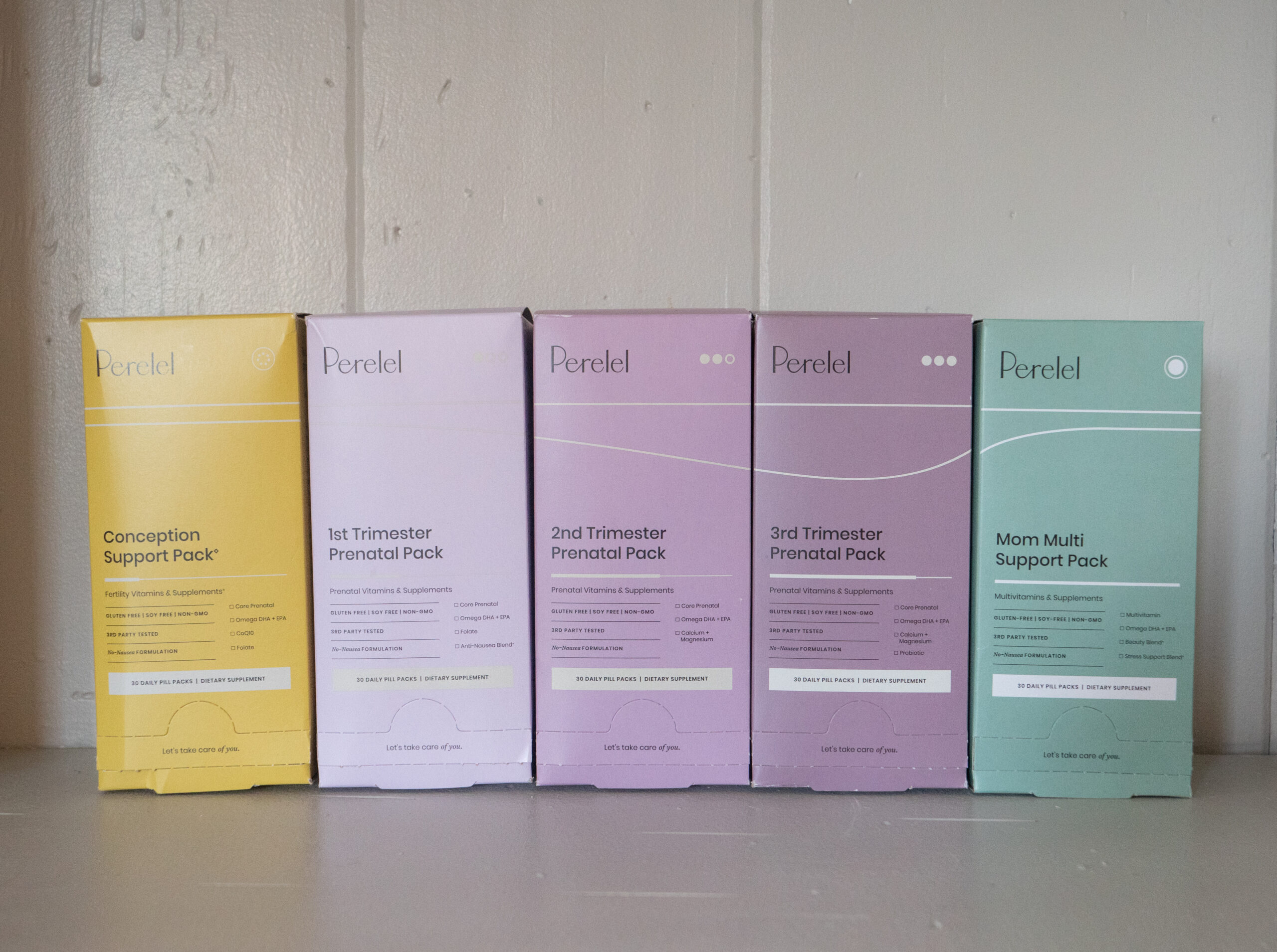 Five Peretel prenatal and conception support packs in various colors are lined up on a shelf against a white wall.