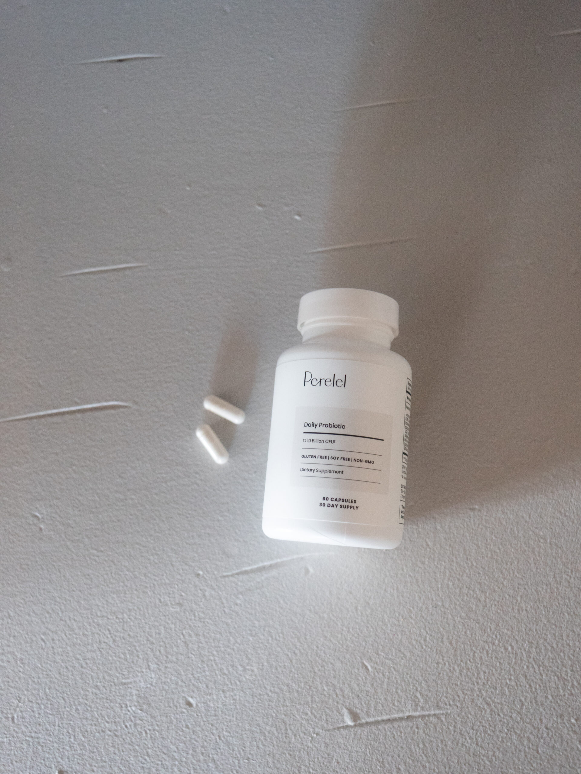 A white bottle labeled "Daily Probiotic" with two white capsules next to it on a textured gray surface.