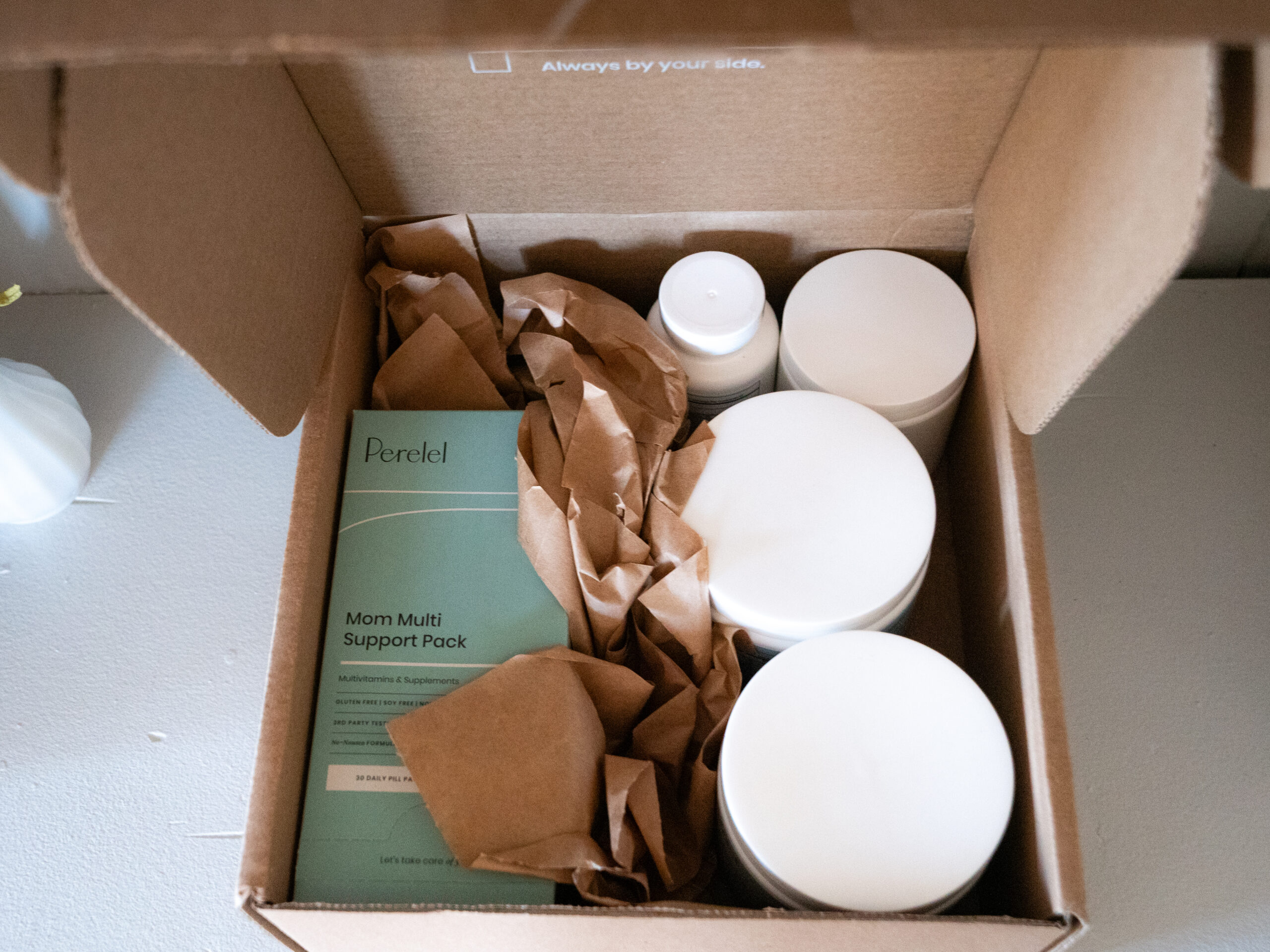 Open cardboard box containing bottles and a green packet labeled "Mom Multi Support Pack," surrounded by brown paper.