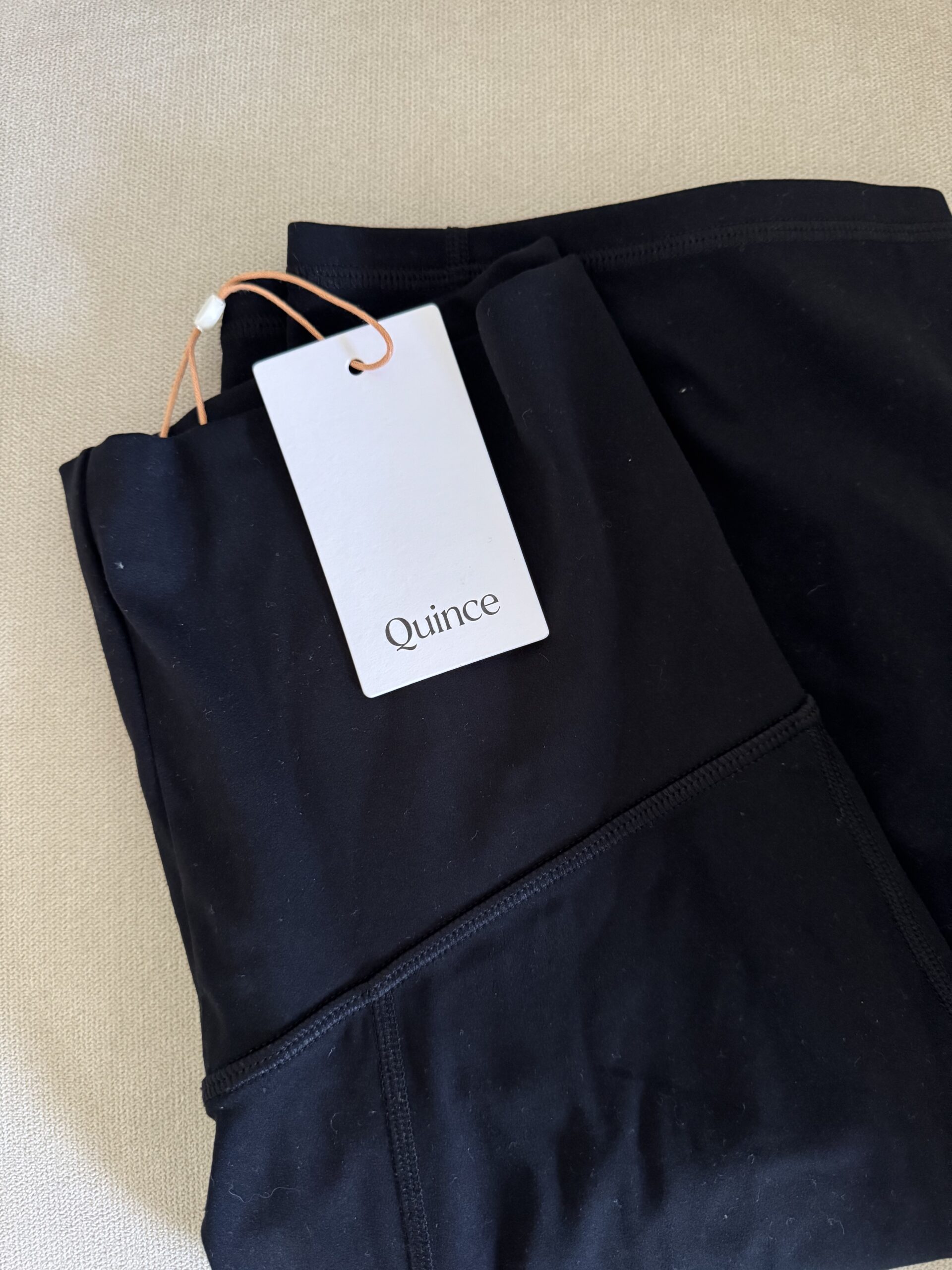 A pair of folded black leggings with a white Quince brand tag attached, placed on a beige surface.