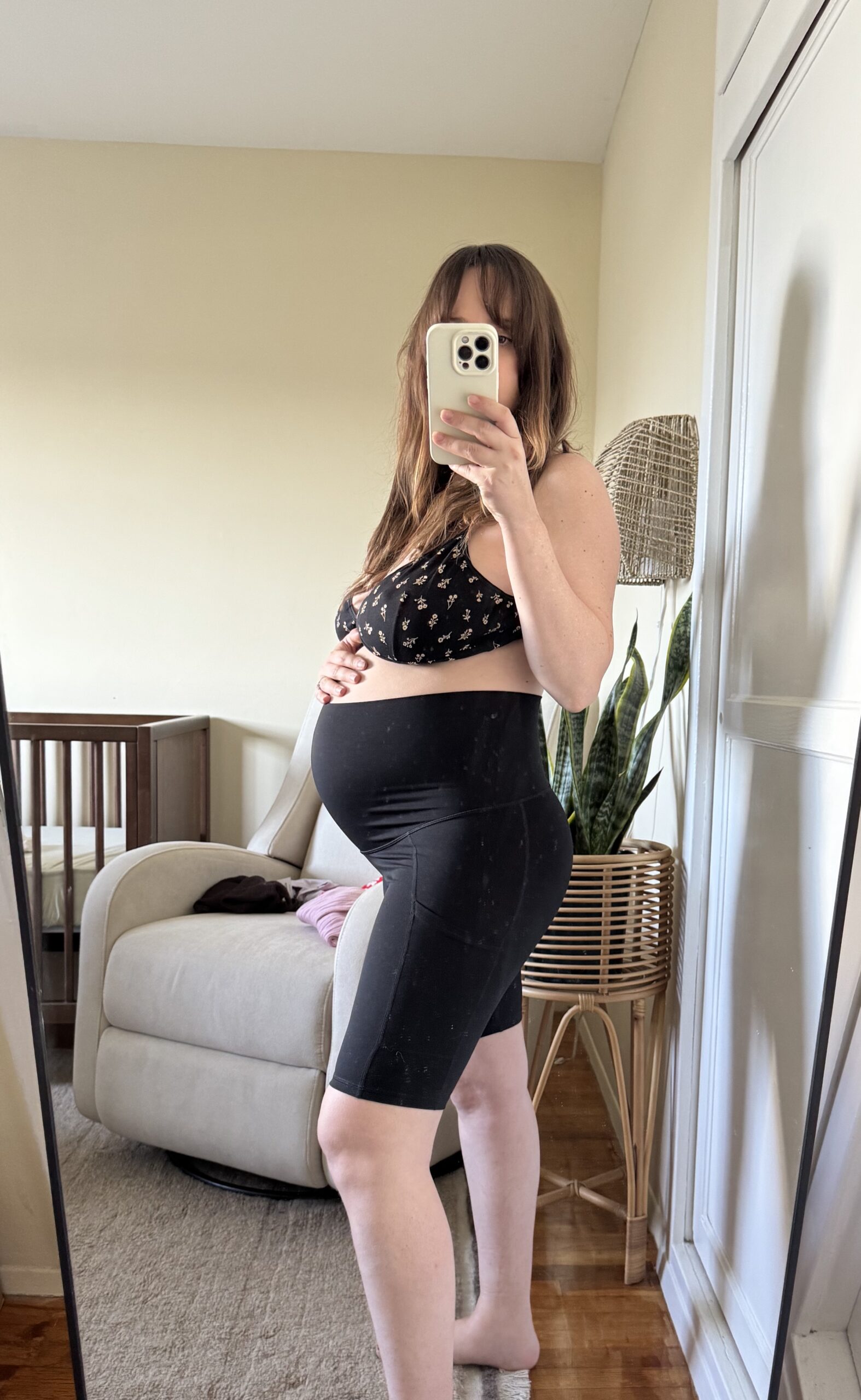 A pregnant woman takes a mirror selfie in a bedroom, wearing a sports bra and biker shorts, with one hand on her belly. A chair, crib, and plant are visible in the background.