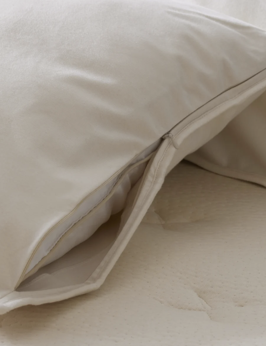 Close-up of an open pillowcase, revealing a partially visible pillow inside. The pillowcase and pillow are both light-colored, resting on a similarly colored fabric surface.