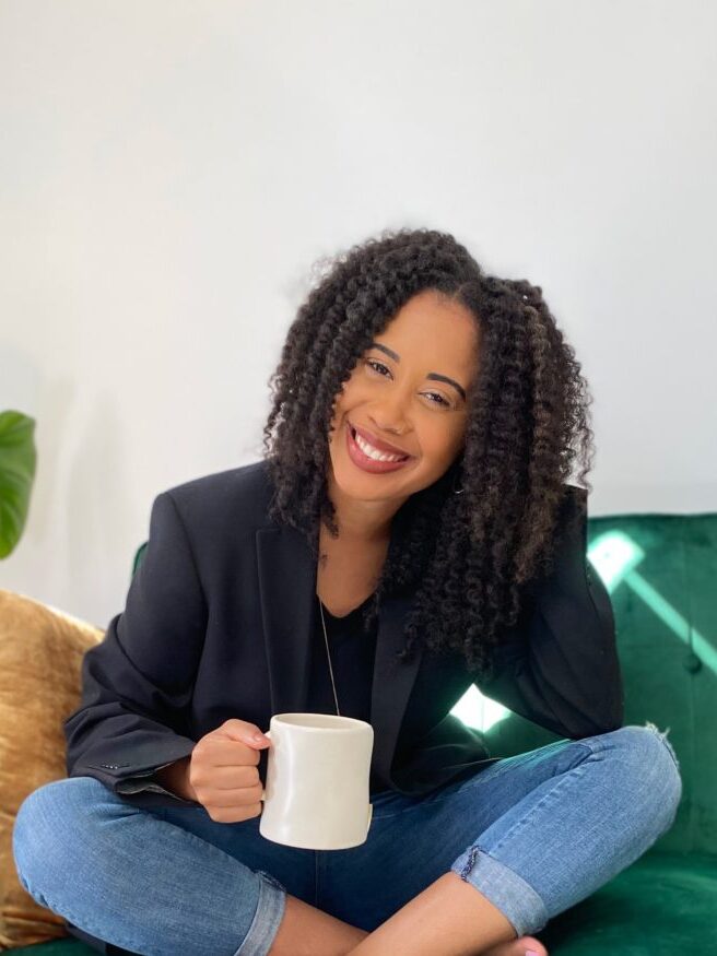 A person with curly hair, wearing a black blazer and jeans, sits cross-legged on a green couch, smiling while holding a white mug.
