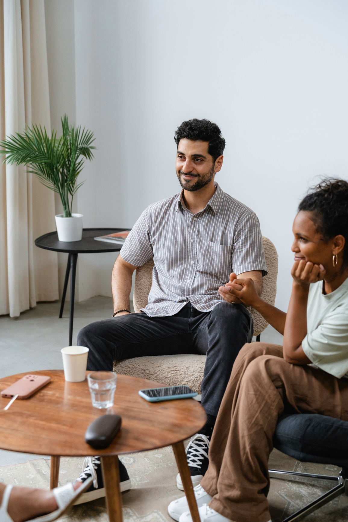 A man and a woman sitting in a living room, engaged in conversation. A small coffee table with a smartphone, glass, and cup is in front of them, and a potted plant is in the background.