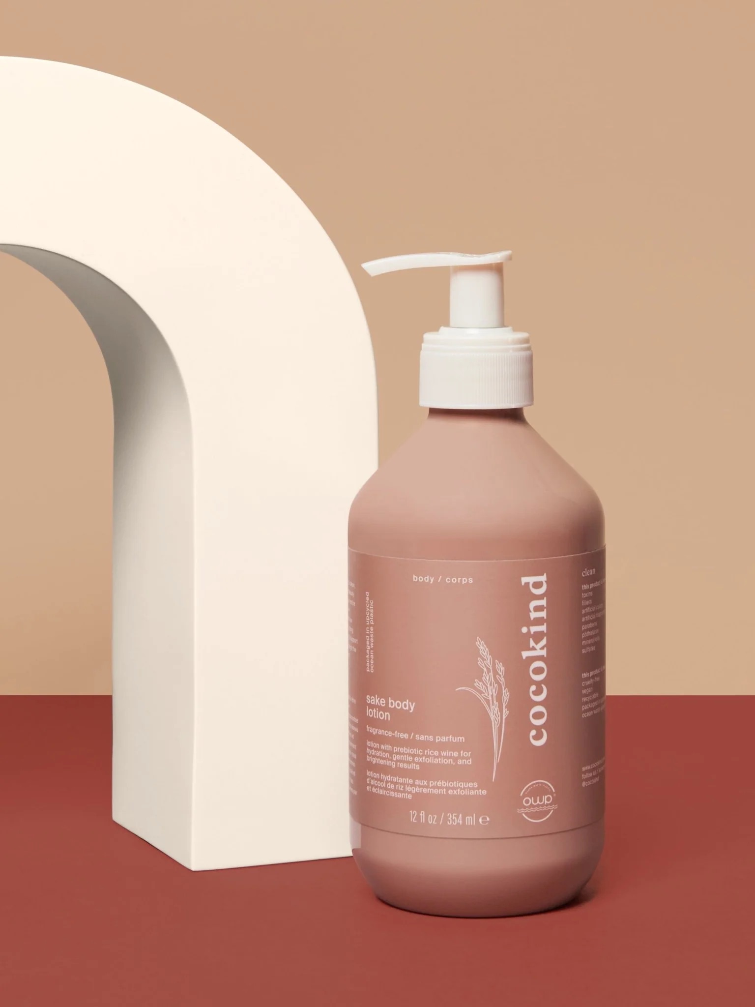 A bottle of Cocokind sake body lotion with a pump sits on a red surface beside a white curved arch, against a beige background.