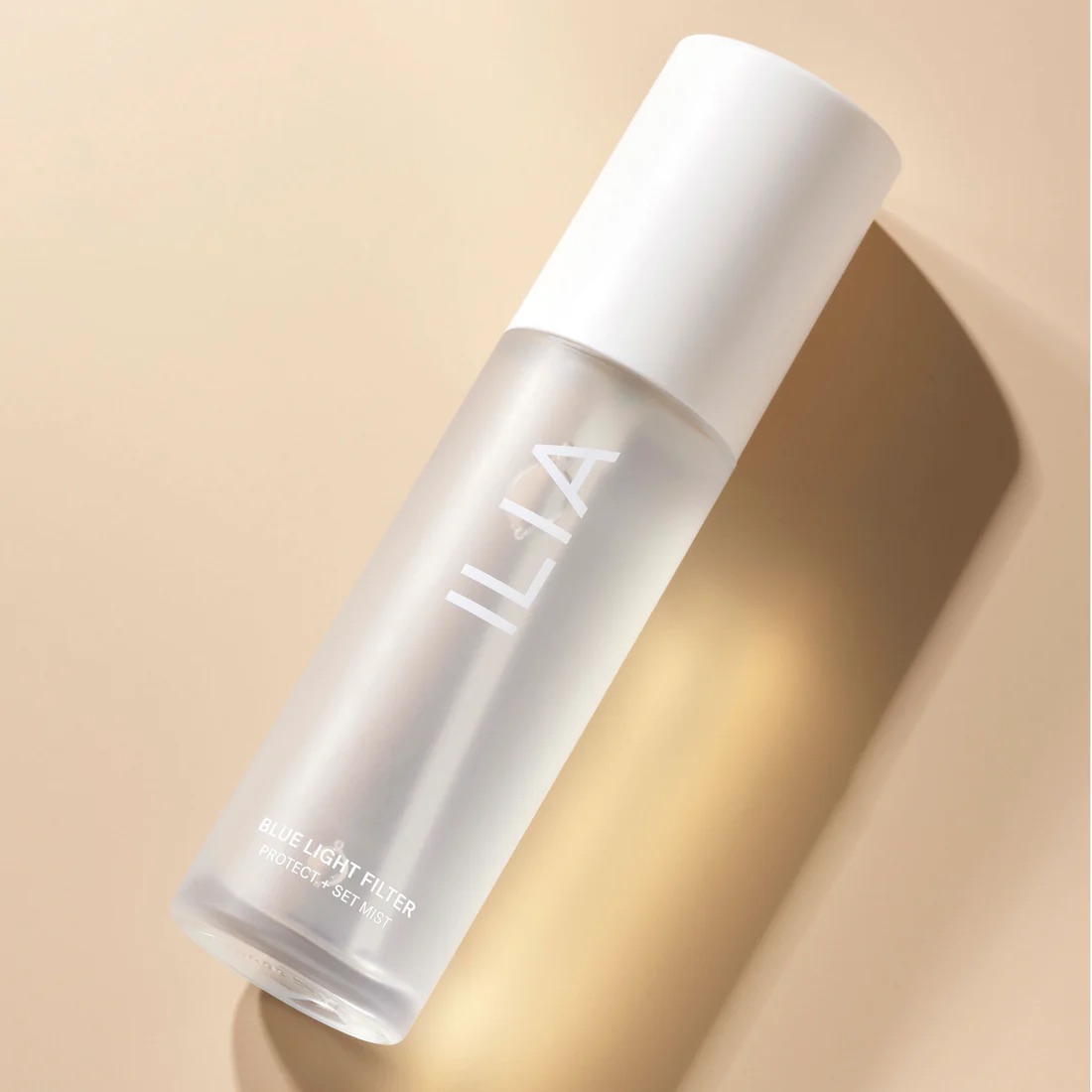 A frosted bottle of ILIA Blue Light Filter Protect + Set Mist with a white cap, lying on a beige surface with a soft shadow.