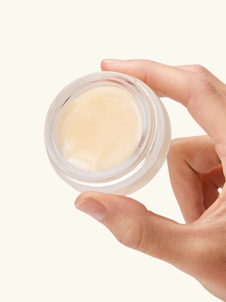 A hand holds a small glass jar containing a light beige cream or balm, pictured against a plain, off-white background.