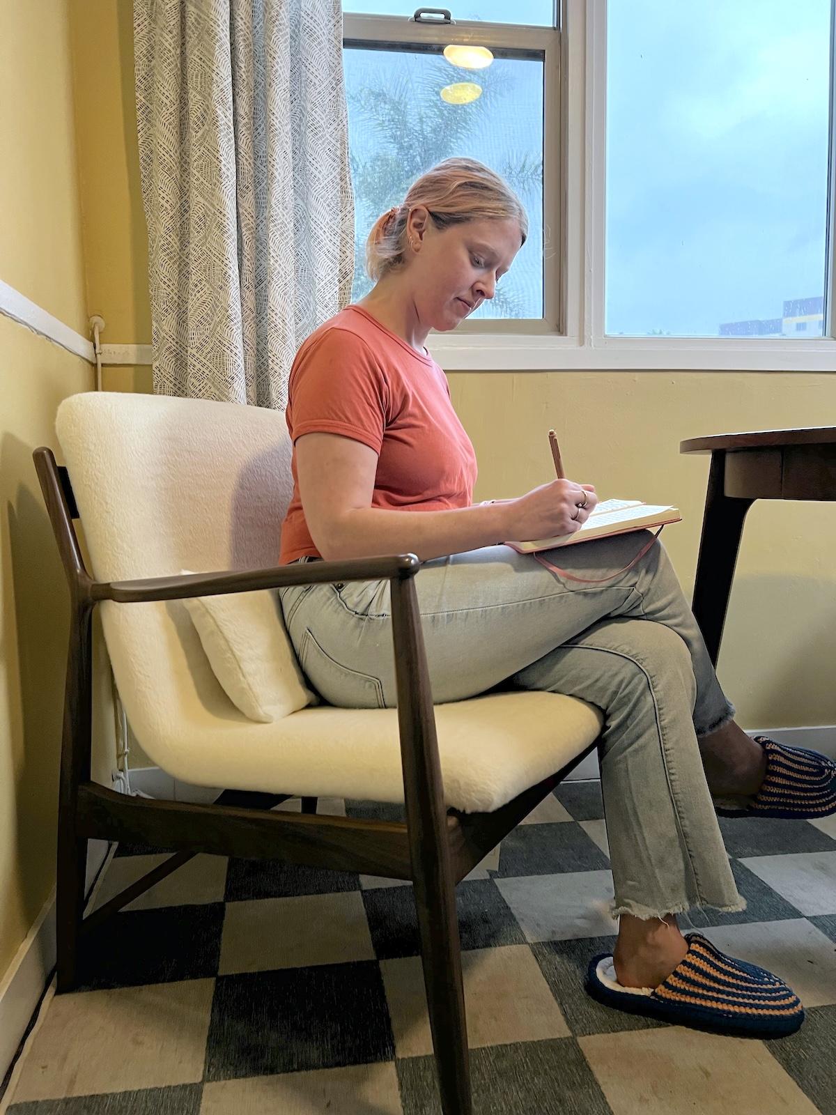 A person in an orange shirt and jeans sits on a chair, writing in a notebook. The room has yellow walls, a window, and a checkered floor.