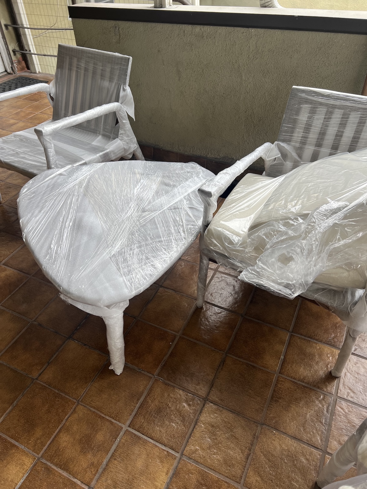 Two patio chairs covered in plastic wrap, placed on a brown tiled floor.
