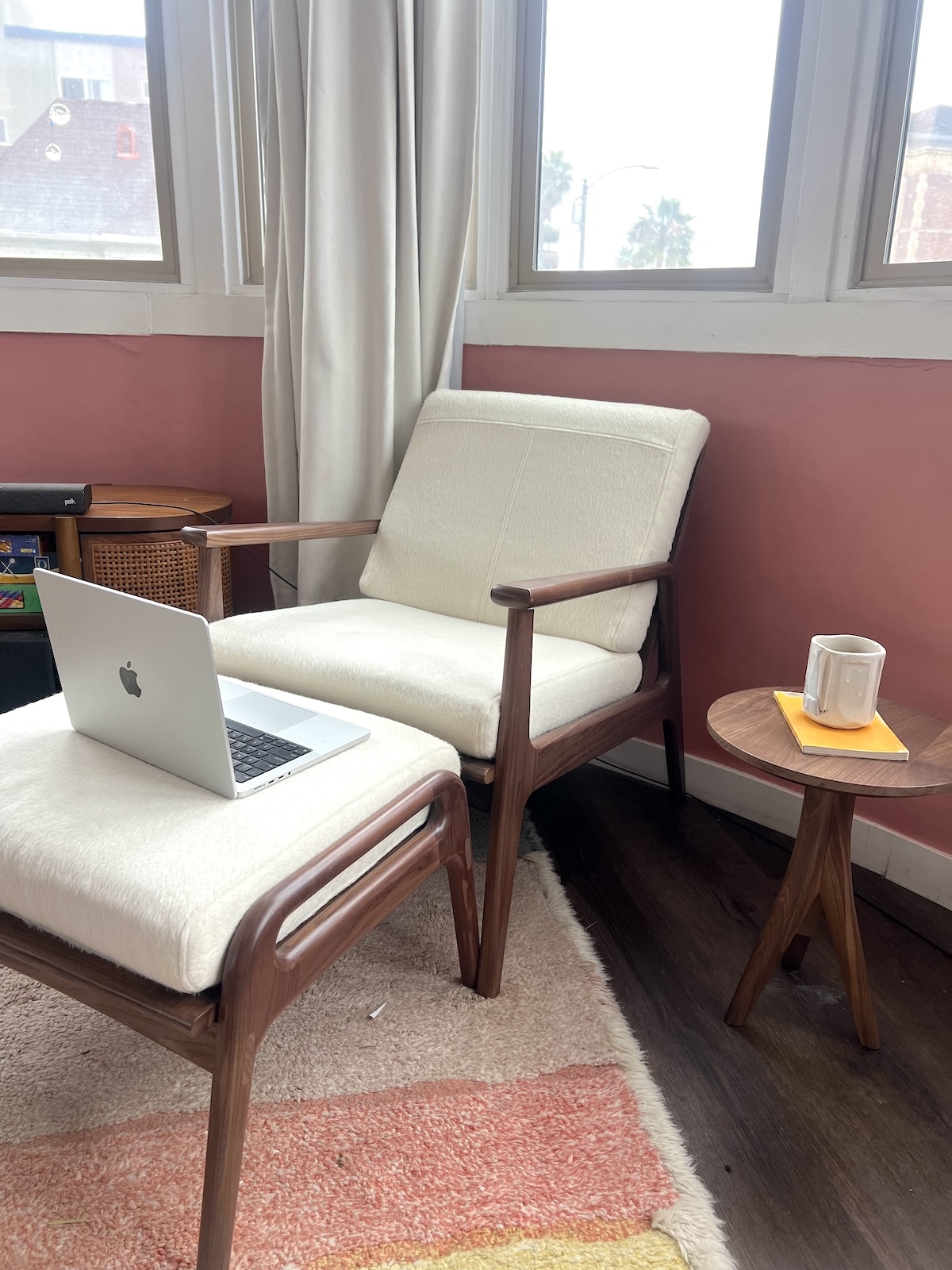 A wooden chair with white cushions holds a laptop. A small round table beside it has a mug on a coaster. There's a window in the background and a pink and beige rug on the floor.