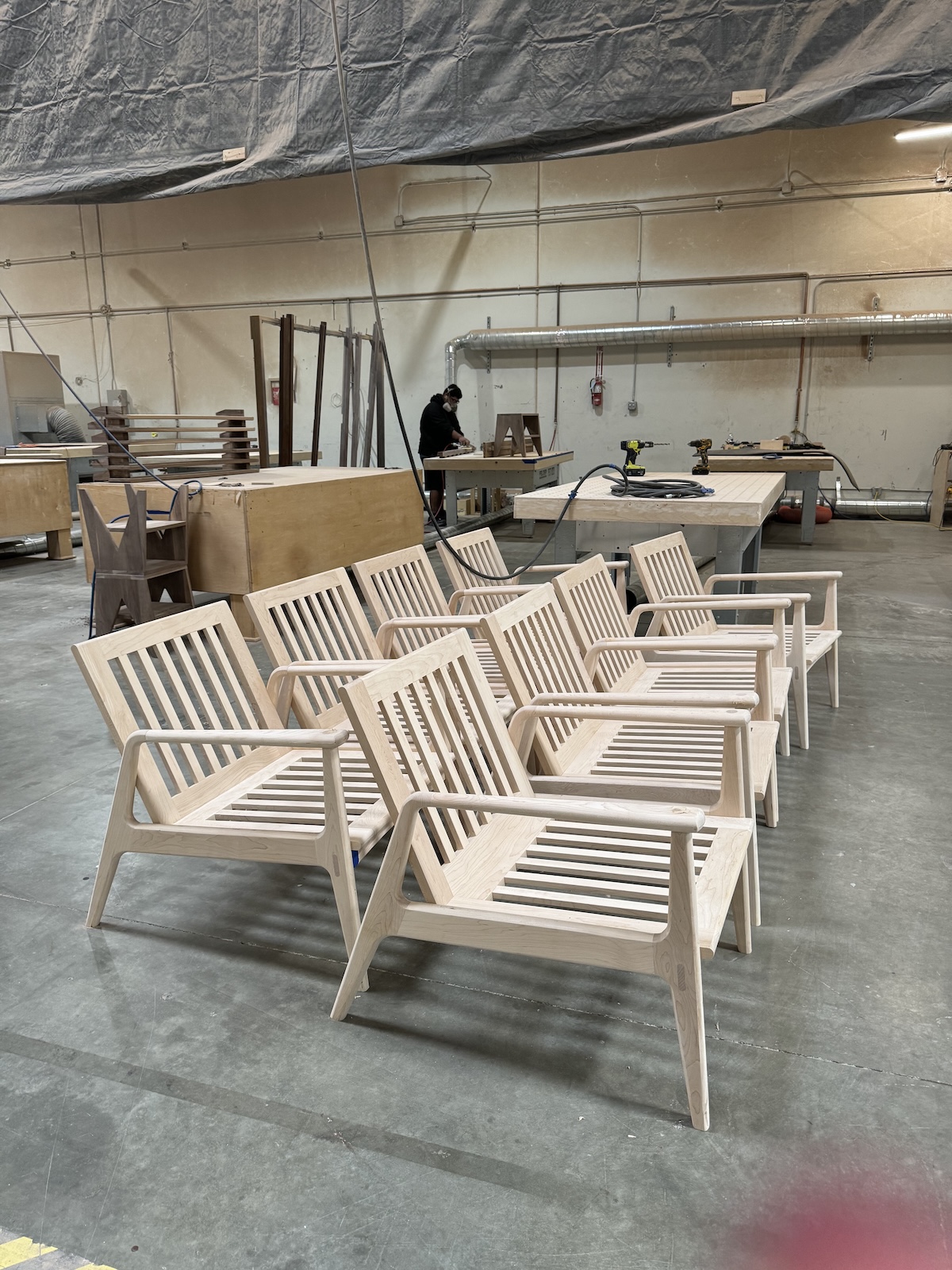 Several unfinished wooden chairs are arranged in rows in a workshop with tools and materials in the background.