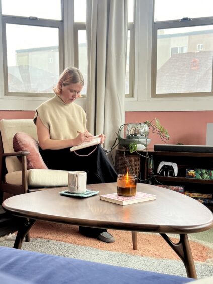 Person sitting on a chair, writing in a notebook. A mug and lit candle are on the table in front. Large windows in the background.