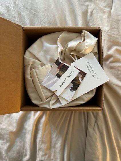 An Avocado brand product wrapped in cream-colored fabric is placed inside an open cardboard box, with tags resting on top.