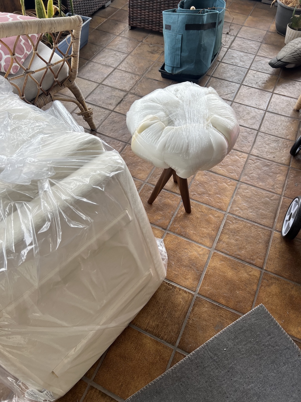 A stool and a boxed item, both wrapped in protective plastic, are placed on a tiled floor near a blue bag and a stroller.