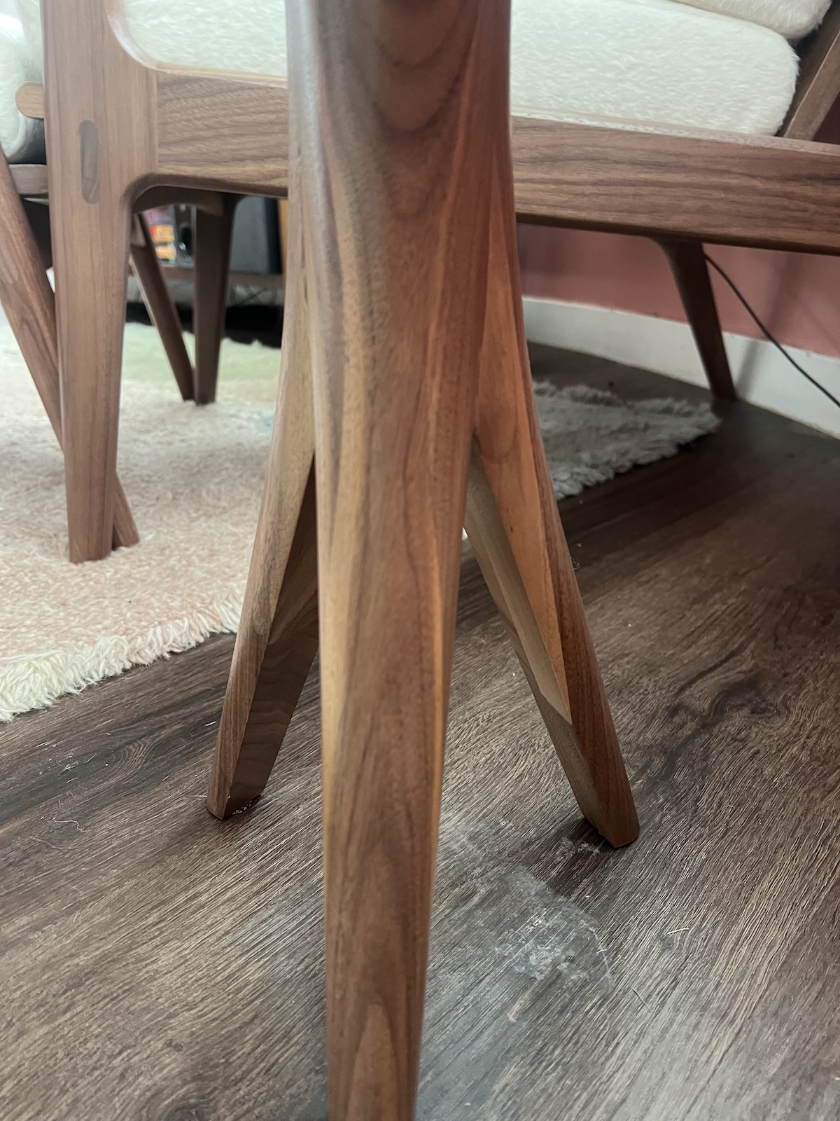 Close-up of a wooden chair leg with a modern design on a wooden floor. Another similar chair is visible in the background.