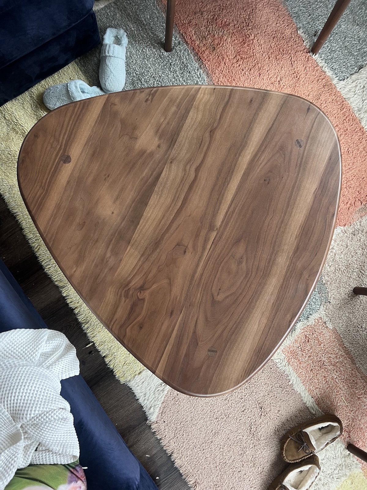 A triangular wooden coffee table on a colorful rug, with slippers and a blanket nearby.