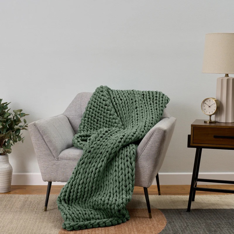 A green chunky knit blanket draped over a gray armchair next to a wooden side table with a lamp and clock, on a multicolored rug.