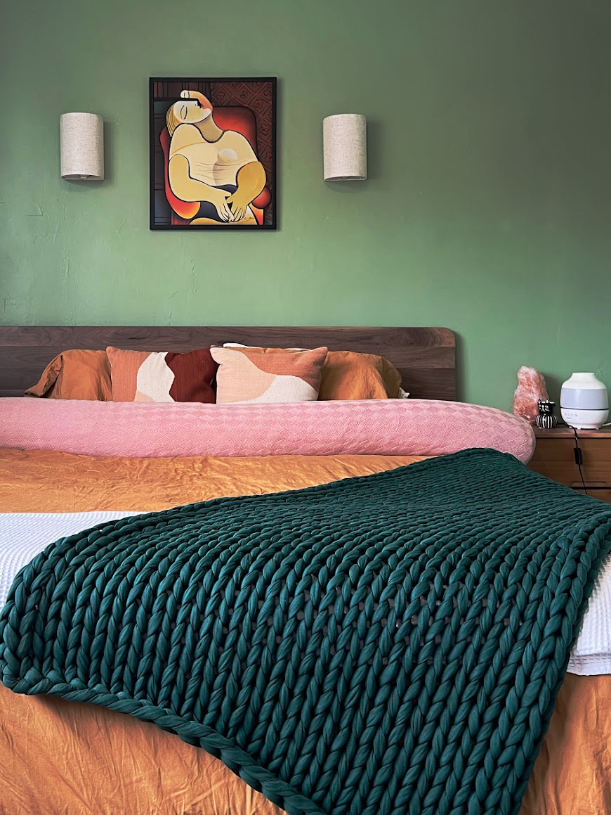 Bedroom with green walls, a large bed with colorful pillows, and a knitted green throw blanket. Artwork of a reclining figure hangs above the bed, with two wall-mounted lights flanking it.
