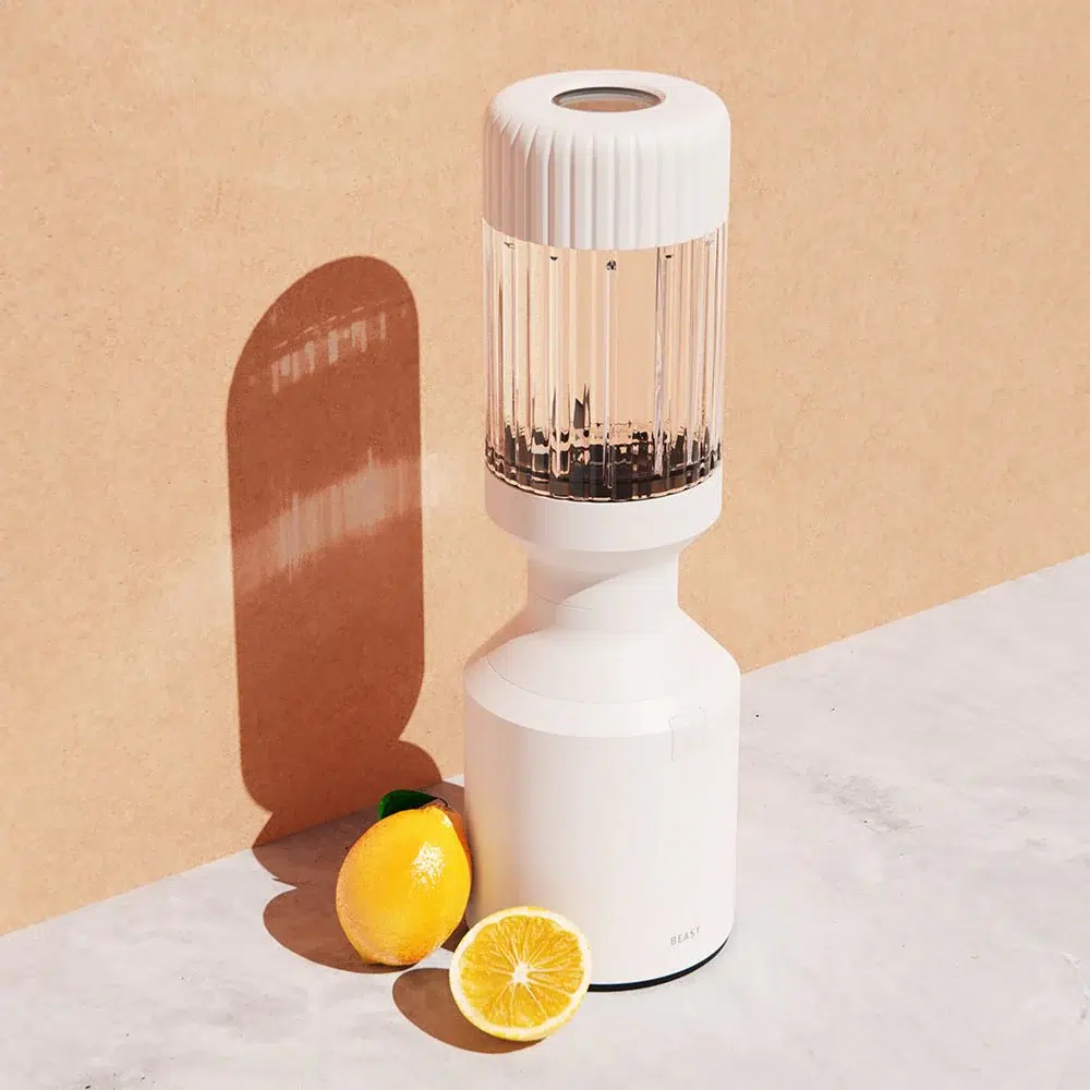 A white blender with a transparent top on a marble surface, next to a whole lemon and a halved lemon, casting a shadow on a beige background.