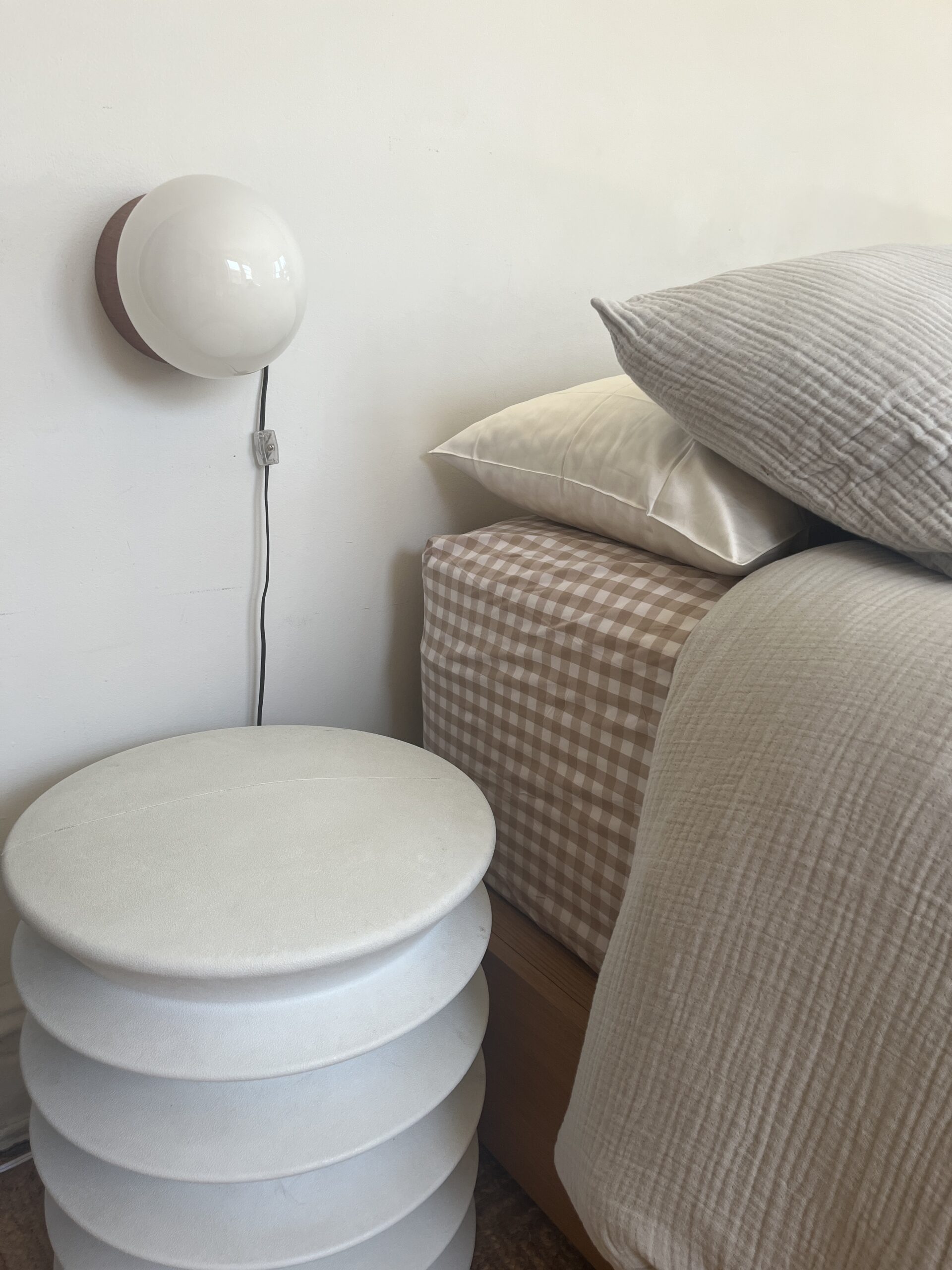 A neatly made bed with gray and checkered bedding is beside a white multi-layered round side table. A wall-mounted spherical lamp is above the table.