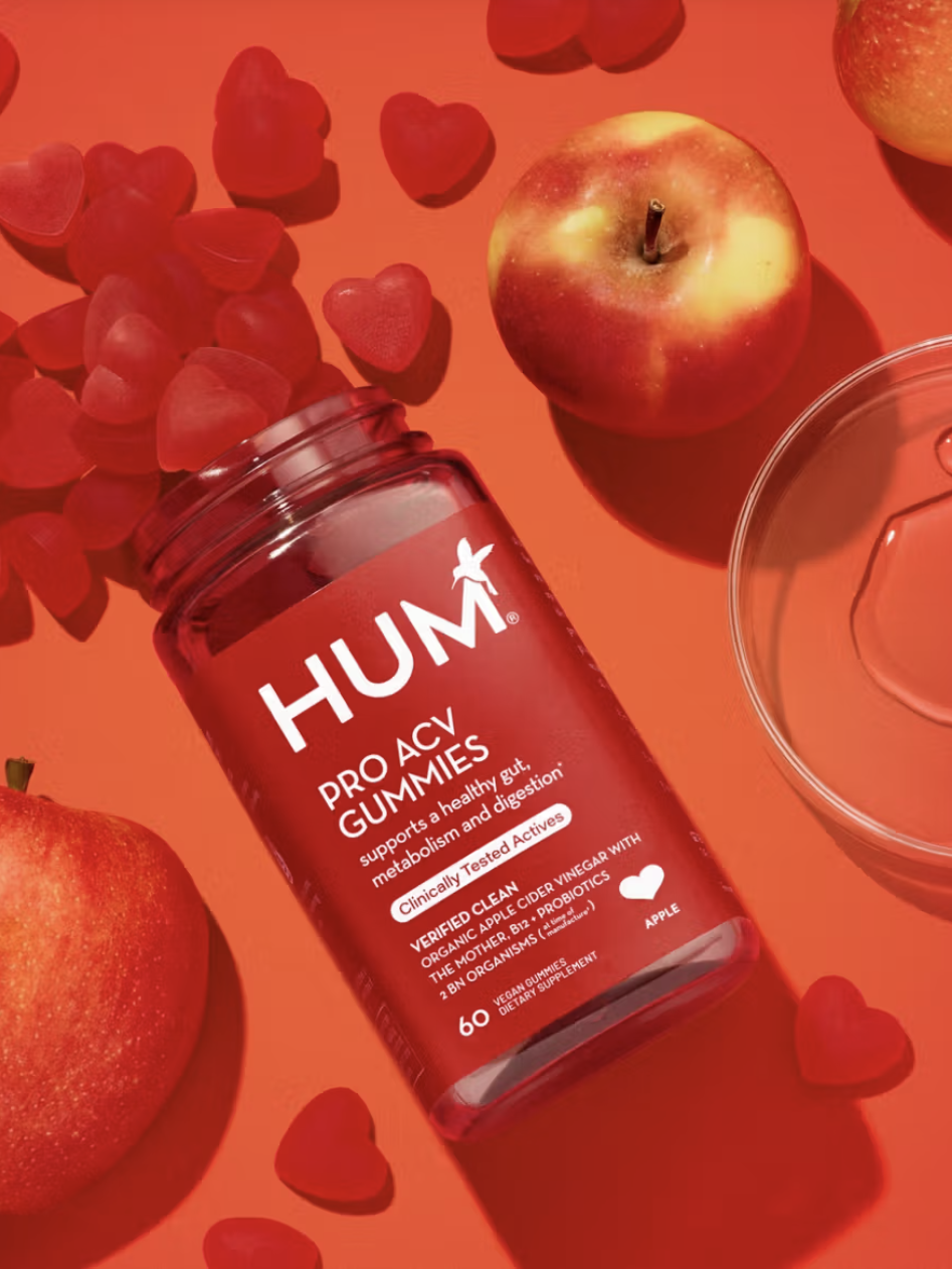 A bottle of HUM Pro ACV gummies is open, surrounded by red apples and a clear plastic lid on a red surface. Heart-shaped gummies are scattered around.
