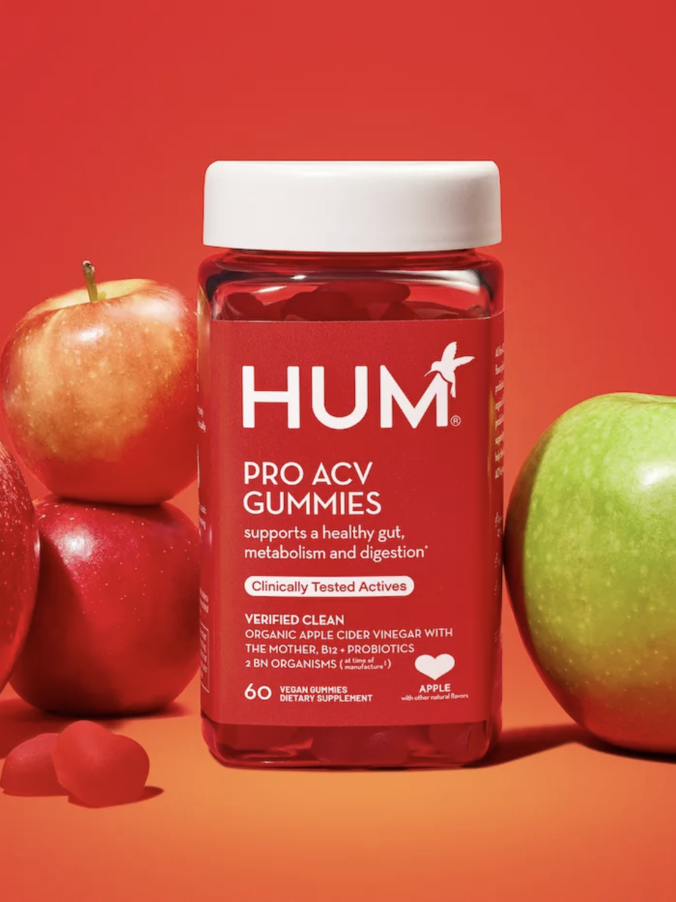 A red jar of HUM Pro ACV Gummies on a red background, surrounded by two red apples and one green apple.