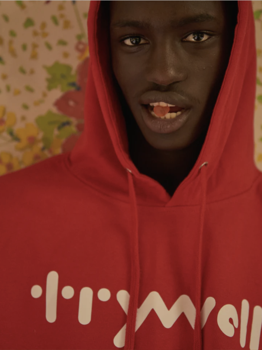 Person wearing a red hoodie with white text, stands against a floral background, holding an orange pill on their tongue.