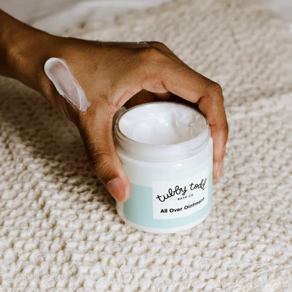 A hand holding an open jar of all over ointment, with some ointment applied on the back of the hand. The jar is placed on a textured beige surface.