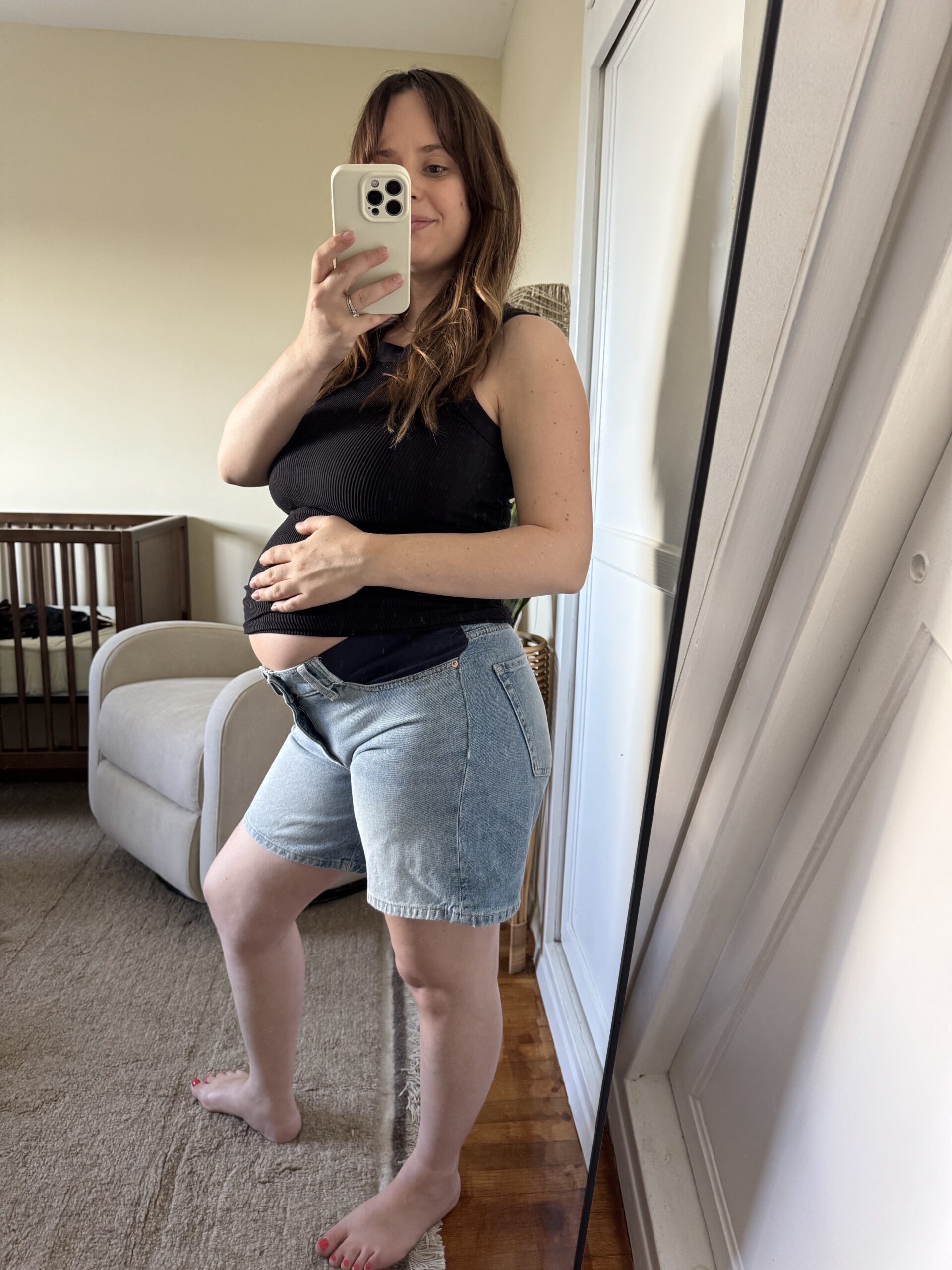 A woman takes a mirror selfie in a bedroom, wearing a black tank top and denim shorts, holding her pregnant belly. A crib and armchair are visible in the background.