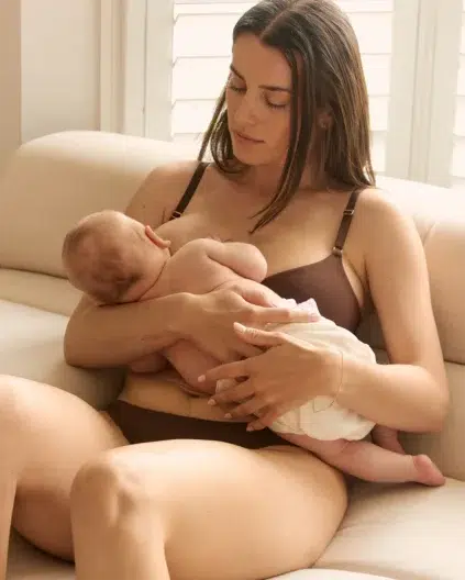 A woman in a brown bra and underwear sits on a beige sofa, breastfeeding a baby.