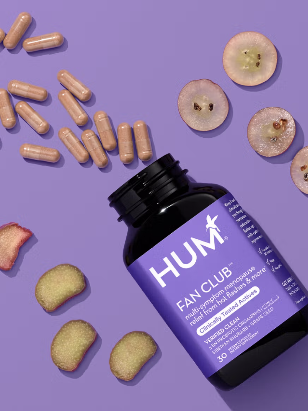 A bottle of HUM Fan Club supplements lies open on a purple surface, surrounded by capsules, grape slices, and pieces of rhubarb.