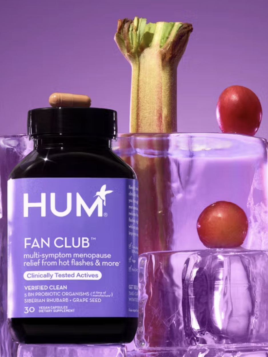 A bottle of HUM Fan Club supplement is placed next to ice blocks containing rhubarb and grapes, set against a purple background.