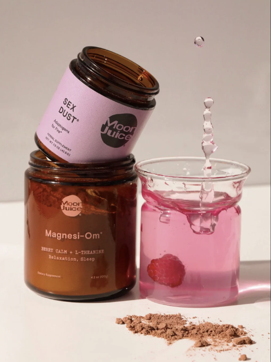 Two jars of Moon Juice supplements, "Sex Dust" and "Magnesi-Om," beside a glass with pink liquid containing a raspberry, and a splash above. Brown powder is scattered on the surface.
