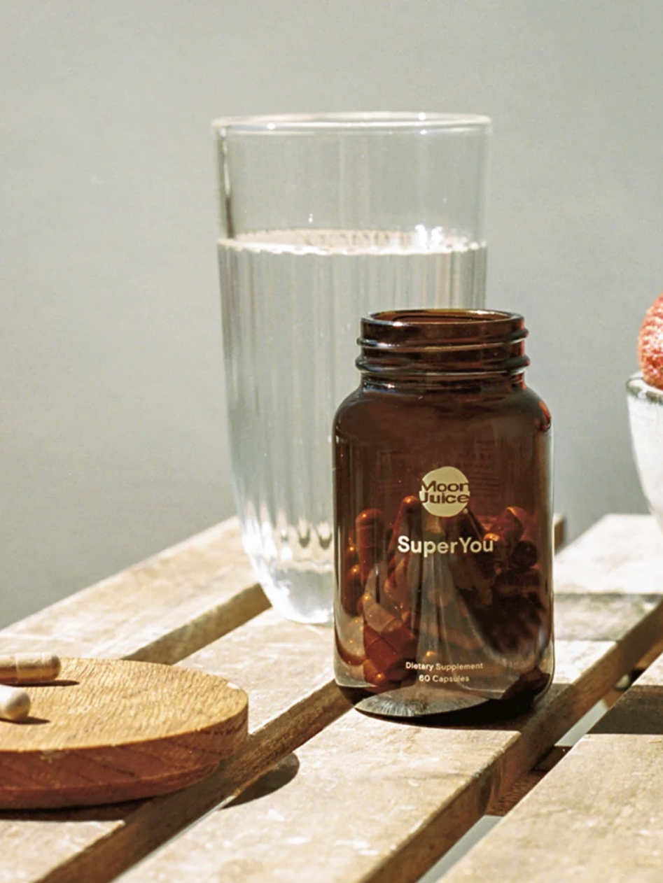 A bottle labeled "SuperYou" on a wooden table with an open cap, two pills, a glass of water, and a bowl of strawberries.