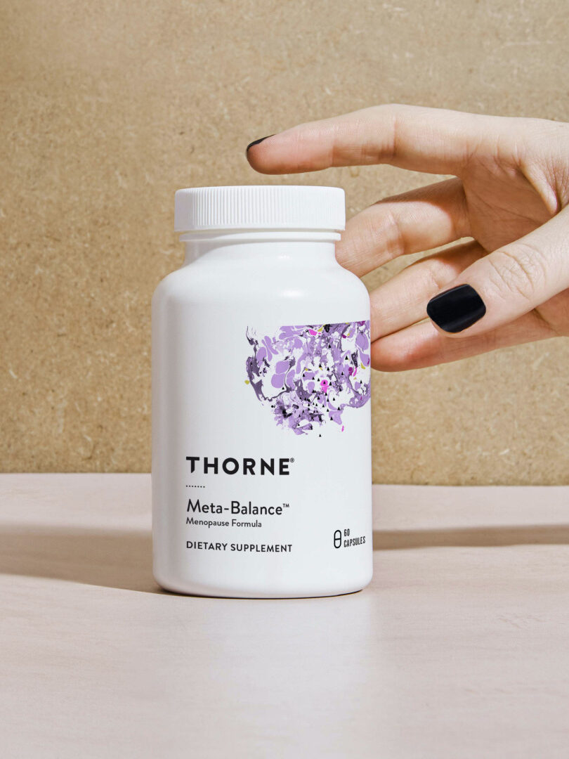 Hand with black nail polish reaching for a white bottle of Thorne Meta-Balance menopause formula dietary supplement, set against a neutral background.
