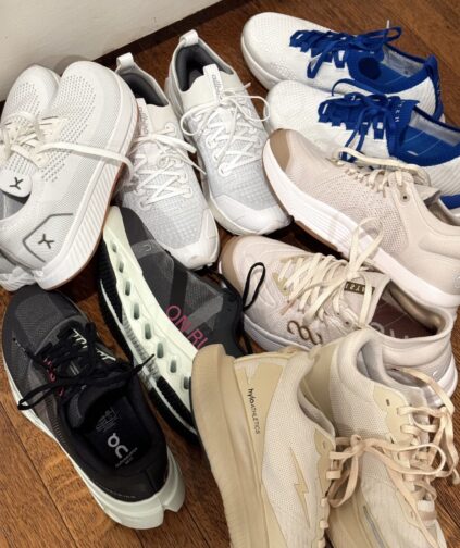 A variety of athletic shoes, mostly in neutral tones with some white and black, arranged on a wooden floor.
