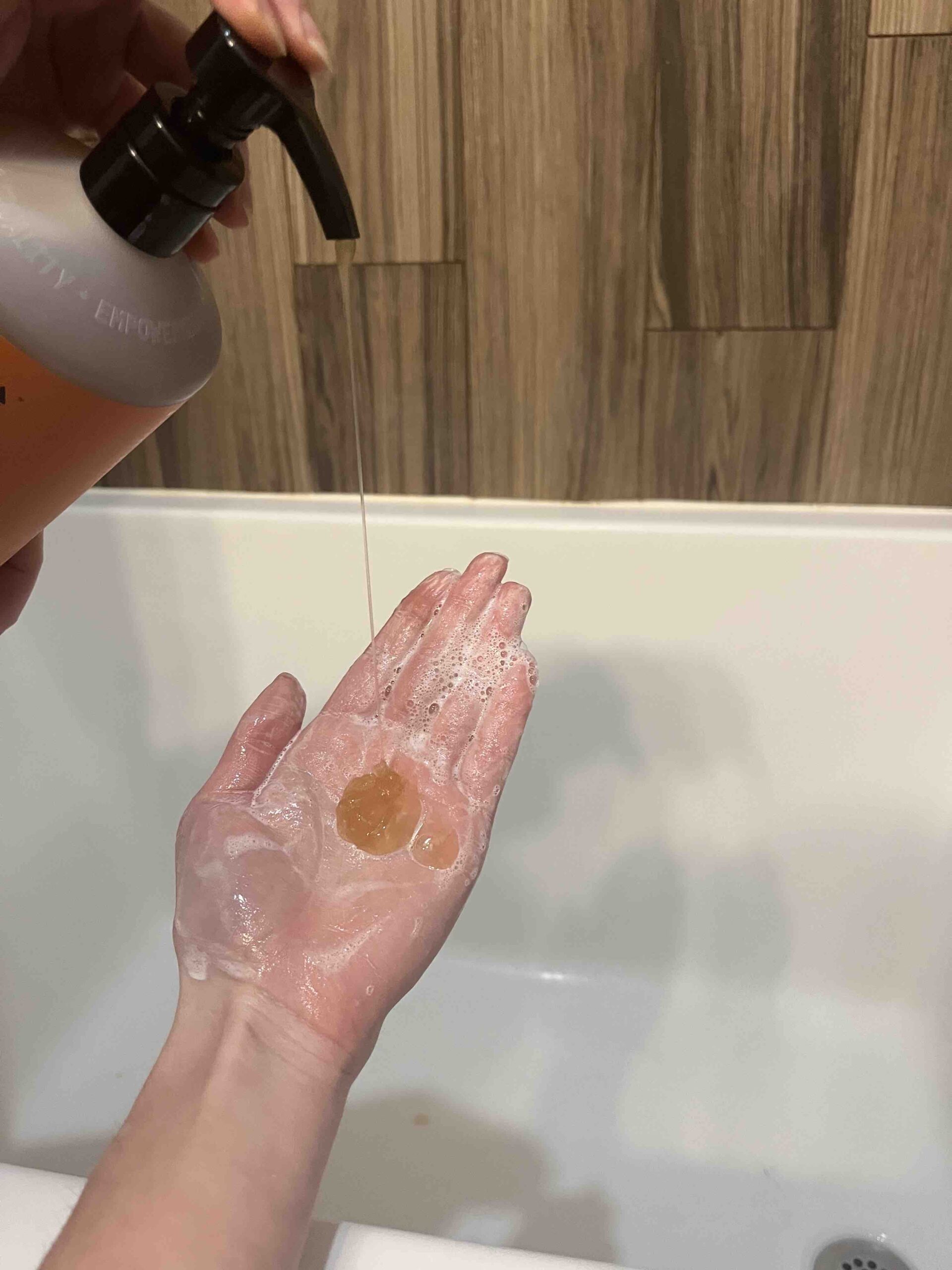 Hand dispensing liquid soap from a pump bottle, creating lather, over a bathtub, with a wooden wall in the background.