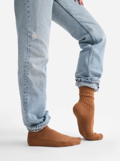 Person wearing light-wash jeans with a small tear and organic brown socks, standing on one foot against a plain background.