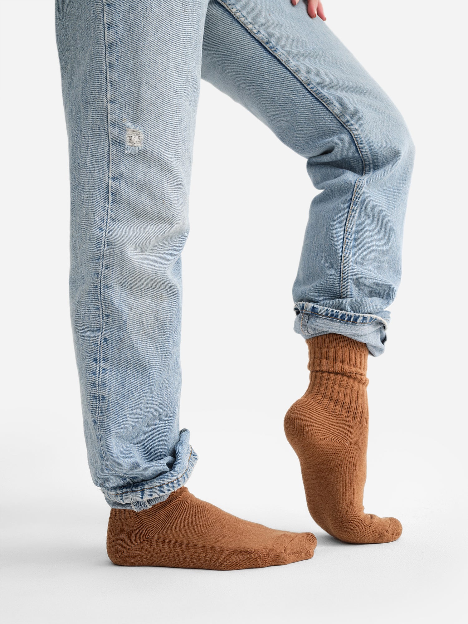 Person wearing light-wash jeans with a small tear and organic brown socks, standing on one foot against a plain background.