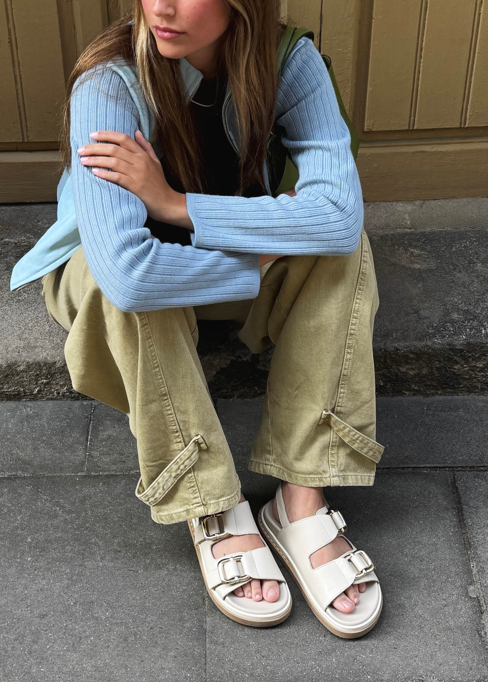A person sits on a stone step wearing light blue ribbed sweater, khaki cargo pants, and beige buckle sandals.